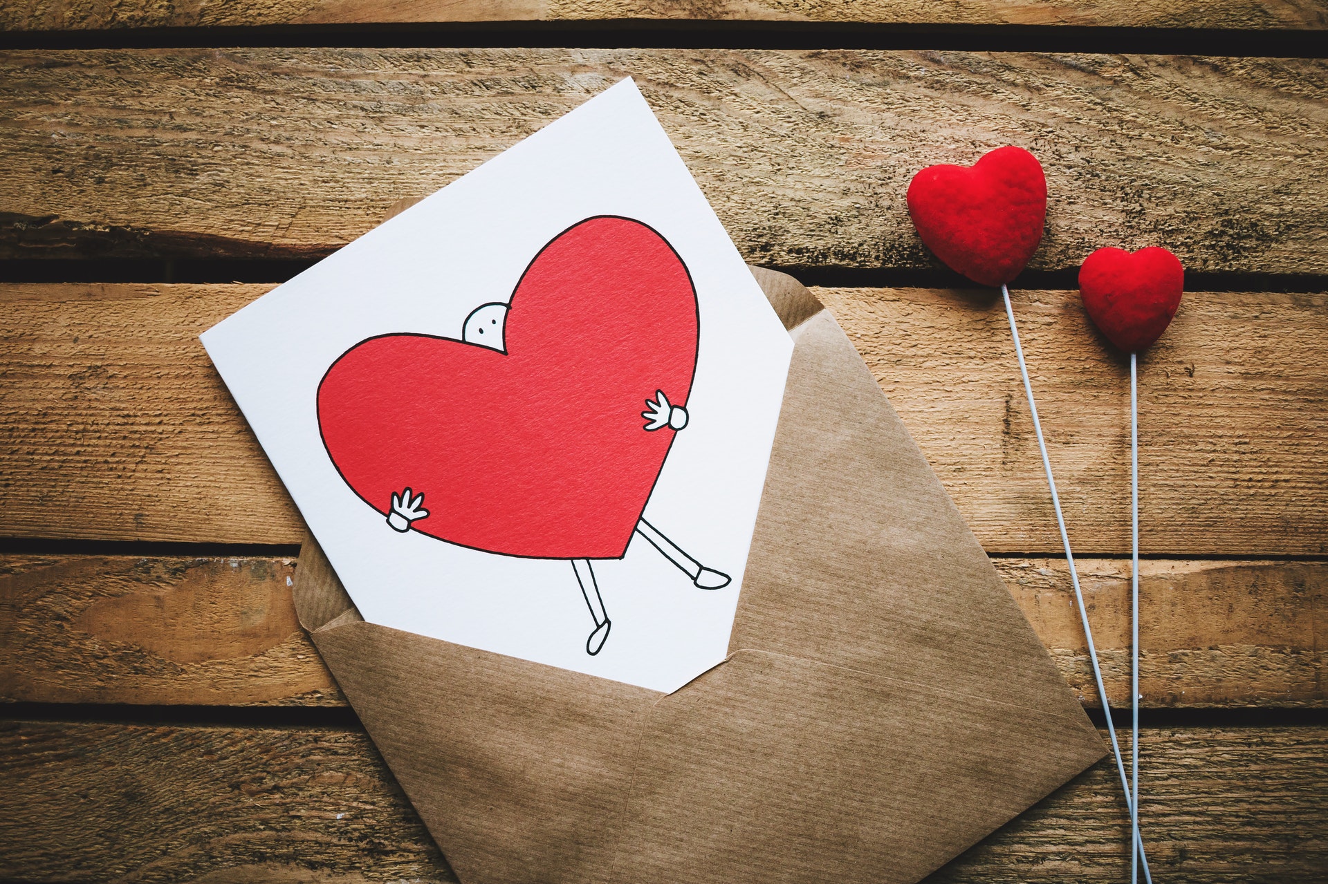 Foto de envelope marrom com cartão branco desenhado com coração e duas hastes com corações vermelhos, tudo sobre superfície de madeira