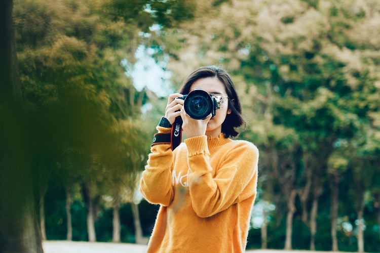 Foto de mulher de blusa amarela segurando uma câmera fotográfica na altura dos olhos