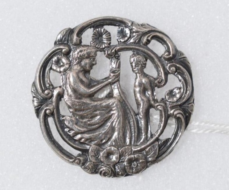 Foto de um botão de prata talhado com ornamentos em estilo rococó, onde se vê a representação do cupido e uma mulher