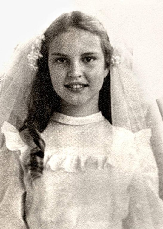 Retrato em preto e branco de Elke Maravilha quando criança, sorrindo, com um vestido branco com babados e um véu branco preso ao cabelo. 