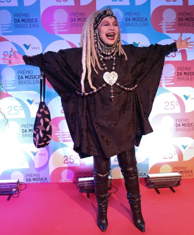 Elke Maravilha posando sorrindo no 25º Prêmio da Música Brasileira em pé e de braços abertos, vestida toda de preto, com botas de cano alto, peruca loira estilo dreadlocks e muitos acessórios. 