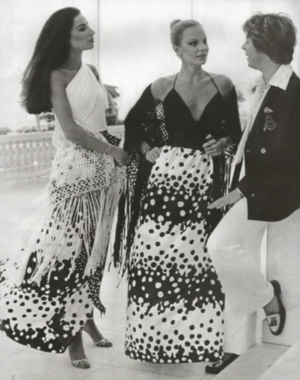 Imagem em preto e branco do estilista brasileiro Guilherme Guimarães jovem em pé a conversar com duas modelos com vestidos longos