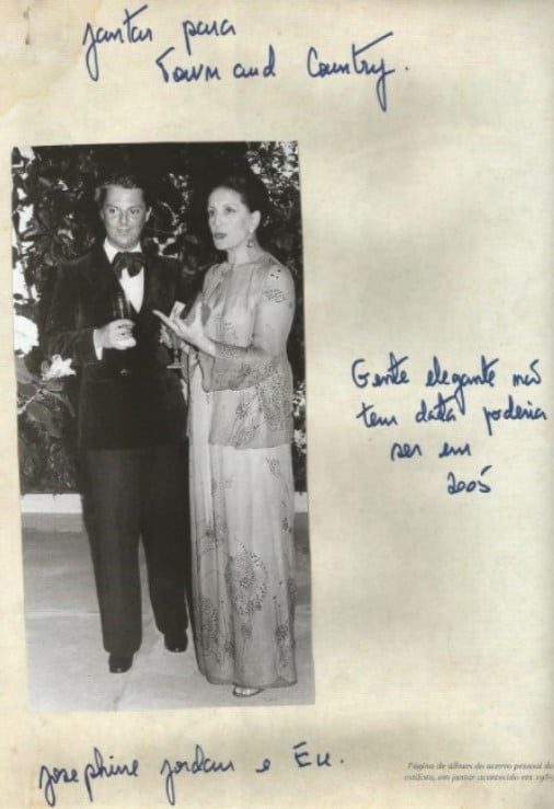 Imagem em preto e branco de Guilherme Guimarães nos anos 80 posando em pé vestido de smoking preto e gravata borboleta preta em um jantar para a Town & Country ao lado de Josephine Jordan