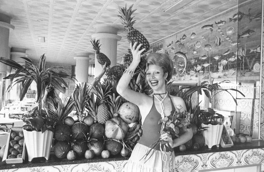 Foto em preto e branco de Elke Maravilha posando com um vestido decotado e segurando um abacaxi na cabeça diante de um espelho rodeado de frutas e plantas.