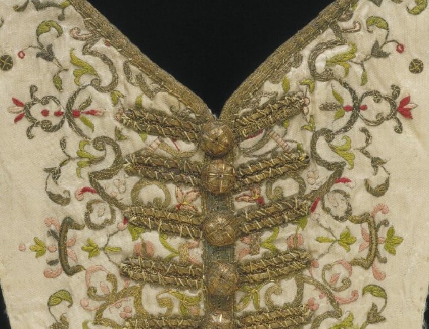 Parte de um corpete femino feito em seda creme com bordados e vários botões do mesmo tecido em fundo preto