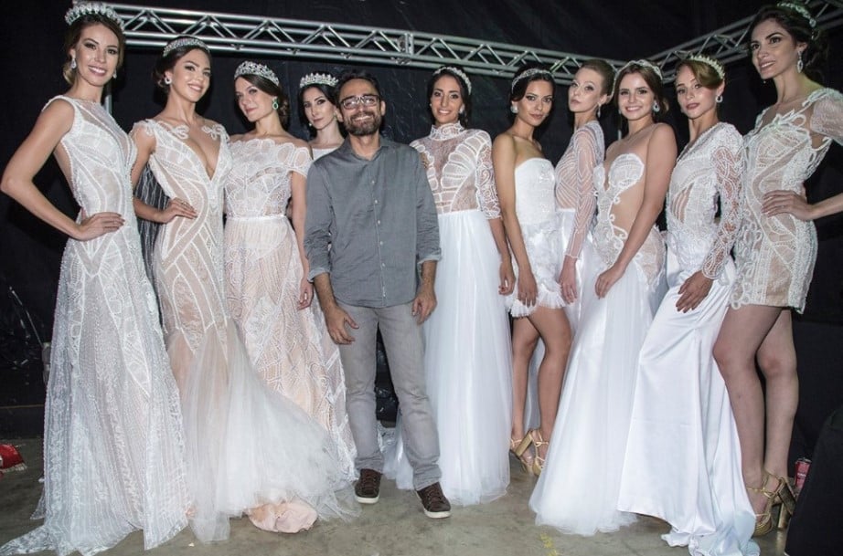O estilista Ivanildo Nunes no centro da foto posando rodeado de modelos trajando vestidos de noiva brancos feitos com rendas e outras técnicas