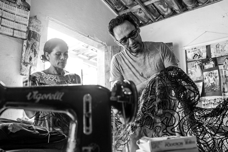 Foto em preto e branco do estilista Ivanildo Nunes a segurar nas mãos uma renda preta ao lado de uma artesã e uma máquina de costura