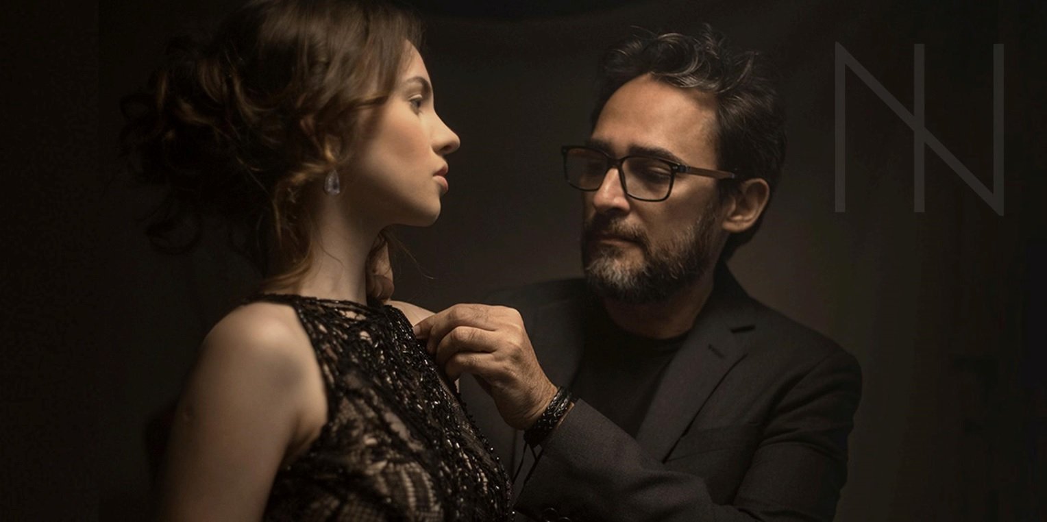 Foto do estilista Ivanildo Nunes posando a ajeitar o vestido de uma modelo, os dois vestidos de preto e em um fundo escuro