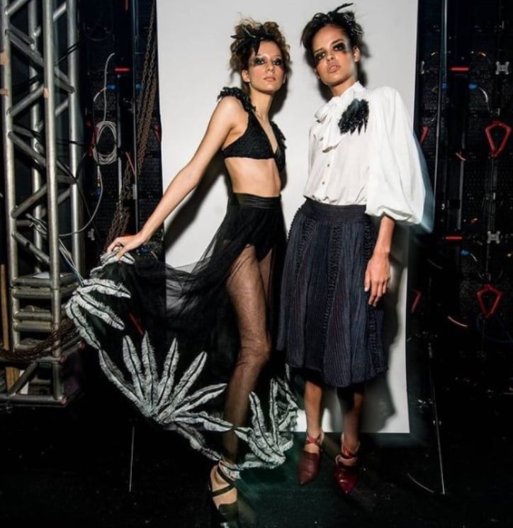 Duas modelos posando em pé com maquiagem preta, uma delas com um top curto e saia transparente preta e a outra com uma camisa branca e saia volumosa preta