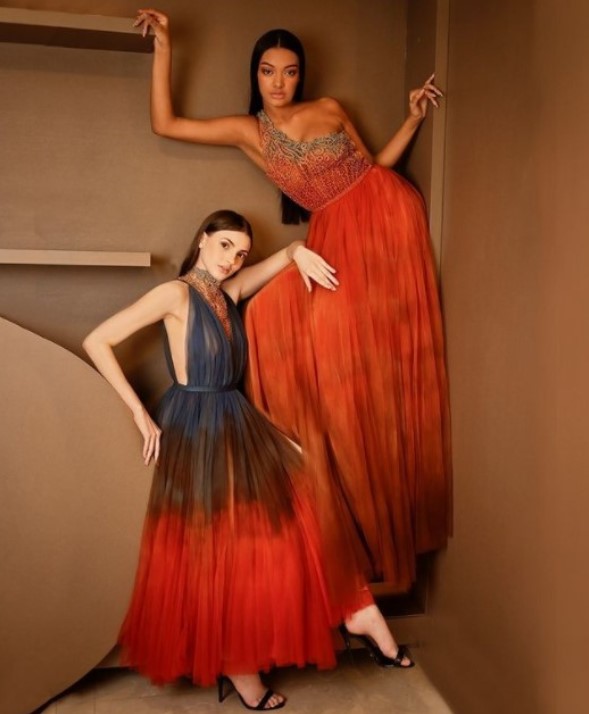 Duas modelos posando contra uma parede com vestidos de festa em tons de laranja e azul do estilista Ivanildo Nunes