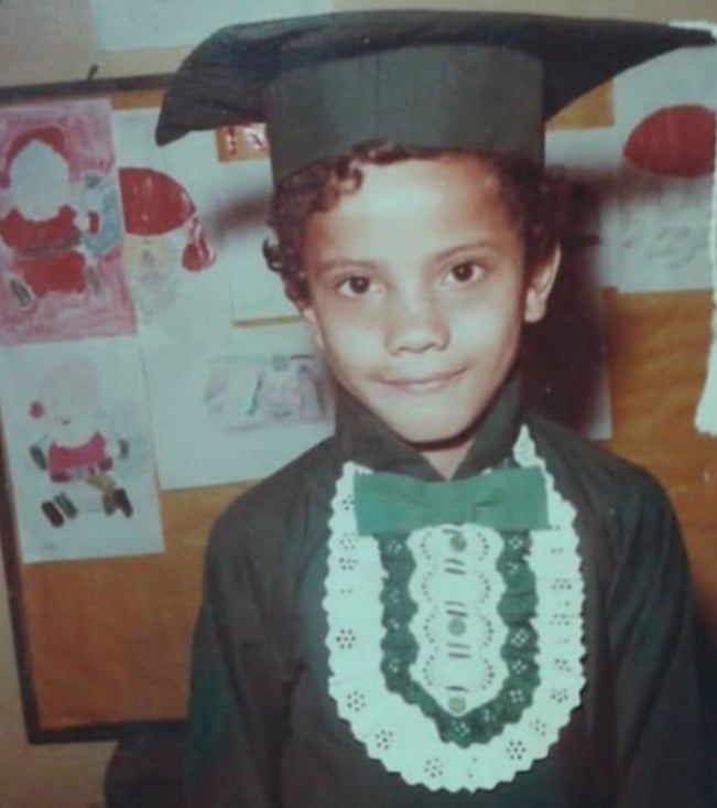Foto colorida de Ronaldo Silvestre criança vestido com um chapéu e roupa de formatura verde diante de um quadro com desenhos pendurados.