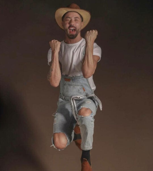 Foto de Rico Melquiades com chápeu de fazendeiro e macacão jeans em pose de comemoração