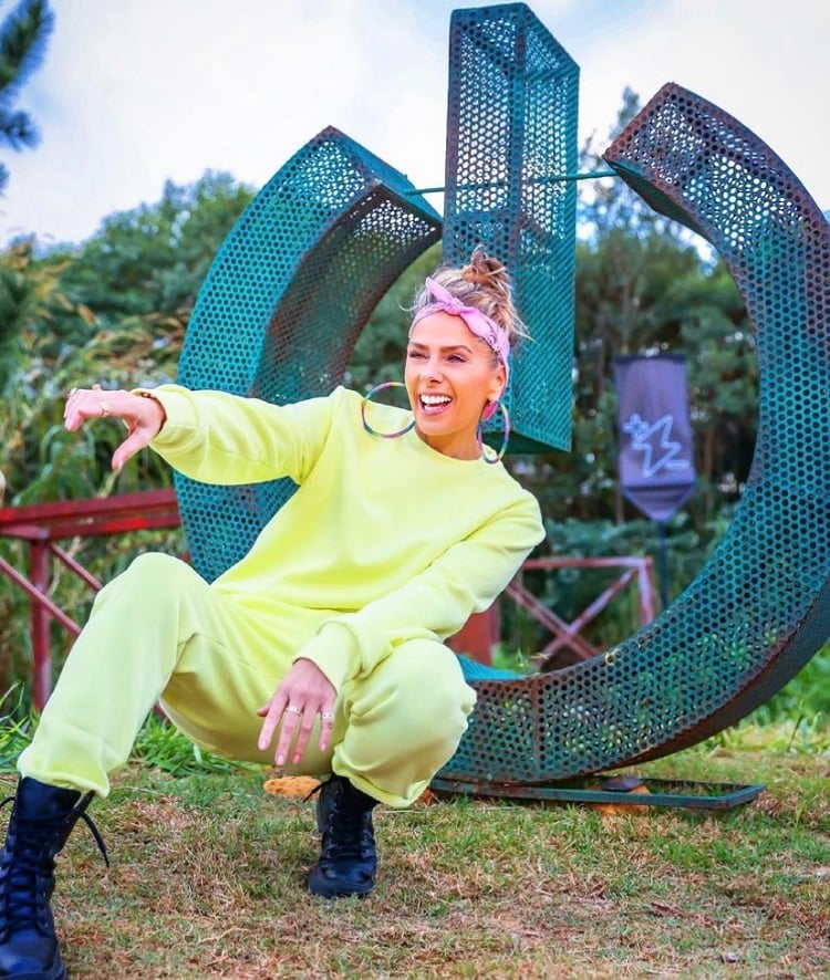 Foto da apresentadora Adriane Galisteu com look neon verde em frente a escultura de ferro em formato da logo do Power Couple