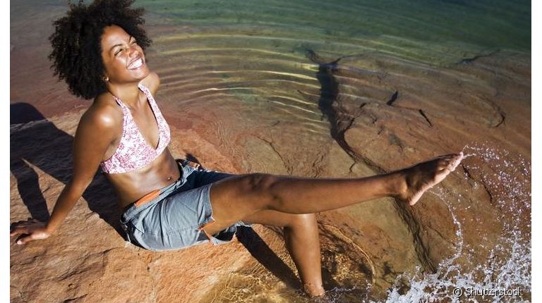 Mulher na beira de um lago sorrindo e demonstrando pernas depiladas