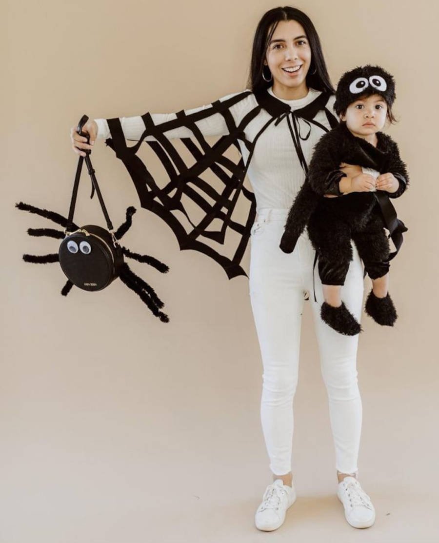 Imagem de uma mãe com capa de teia de aranha segurando sua filha fantasiada de aranha e segurando uma bolsa customizada