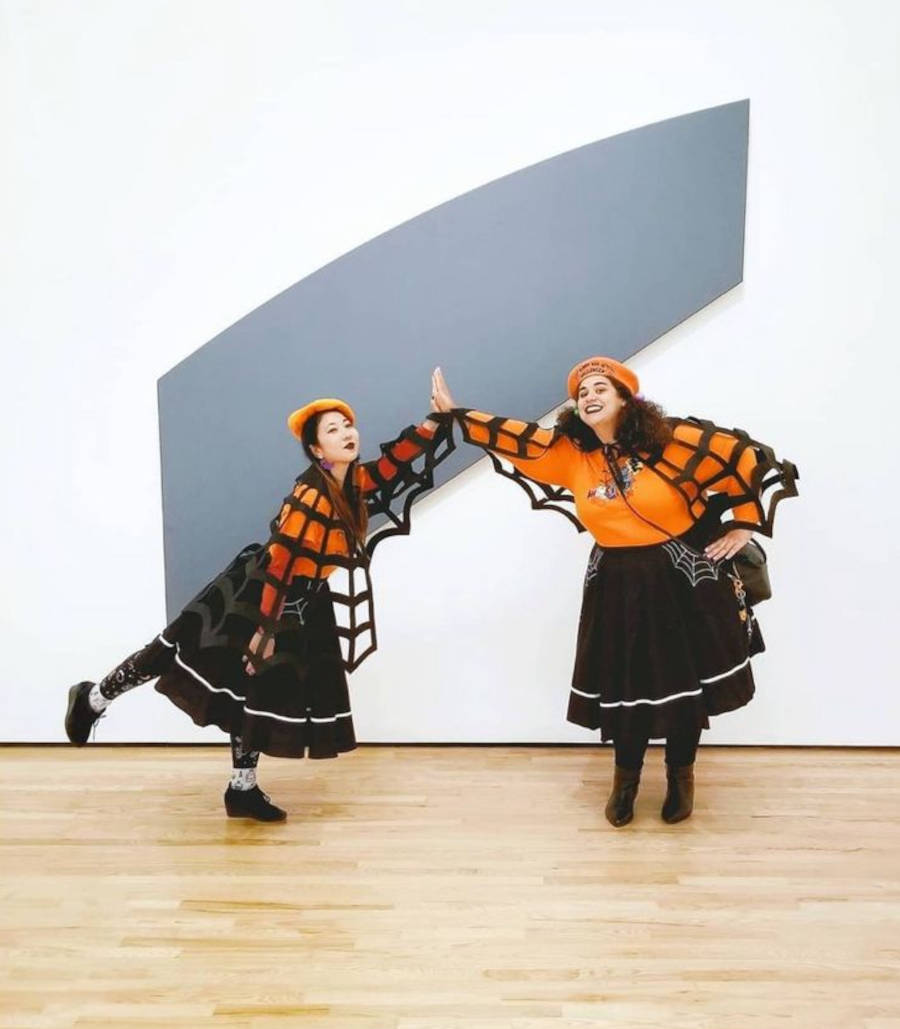 Imagem de duas mulheres distantes encostando as mãos vestidas com fantasia de Halloween composta com a capa de Teia de Aranha