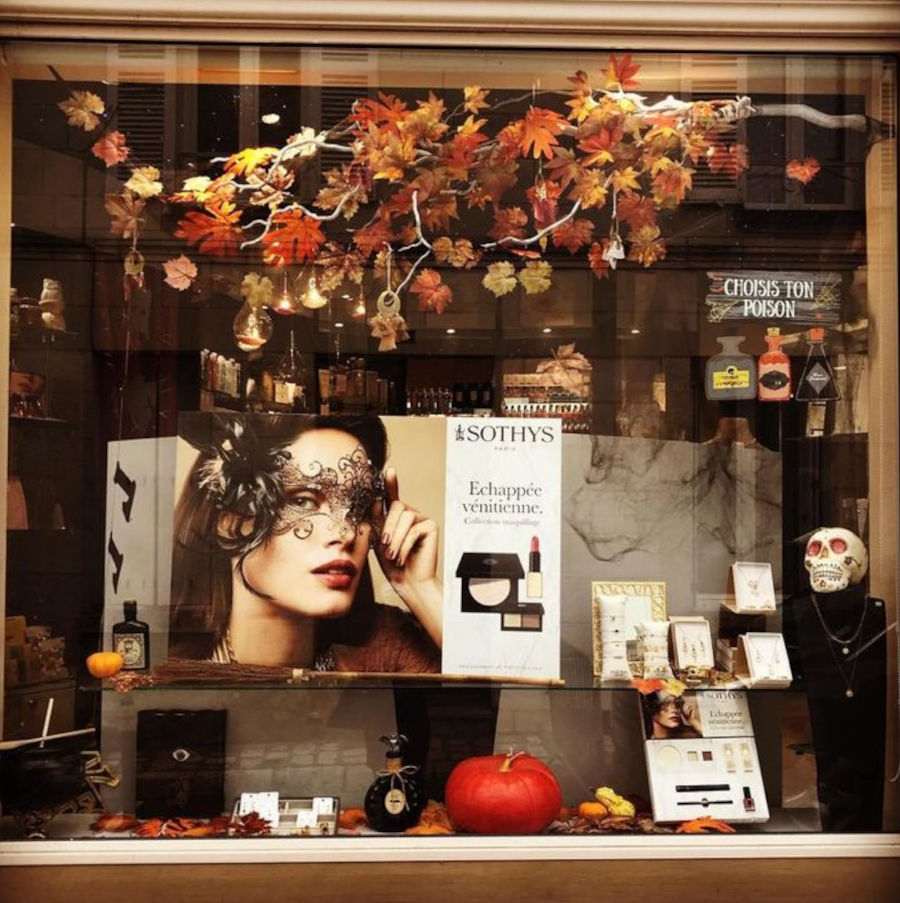 Foto de uma vitrine de loja de cosmético decorada para o Halloween com galhos de árvores laranjas, abóbora, foto de modelo com máscara e caveira