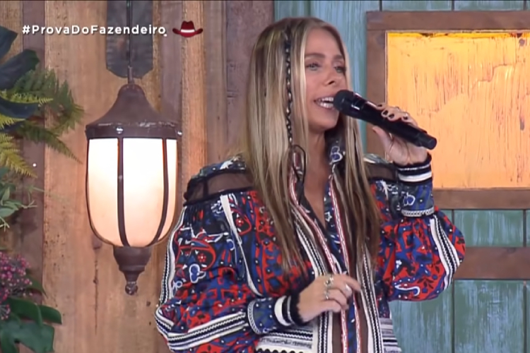 Foto de Adriane Galisteu apresentando A Fazenda. Ela está de cabelo solto e usa blusa colorida. A Fazenda 2021 Programação 06/10
