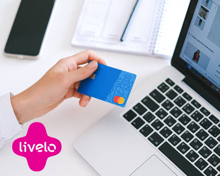 Pessoa fazendo compra online, com notebook, cartão, celular, caderno e lápis. Imagem com logo da empresa Livelo. Foto de pessoa fazendo compras online.