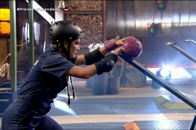 Marina está colocando uma bola em uma gangorra, com macacão azul e capacete. Erasmo, Rico e Solange estão na Roça.