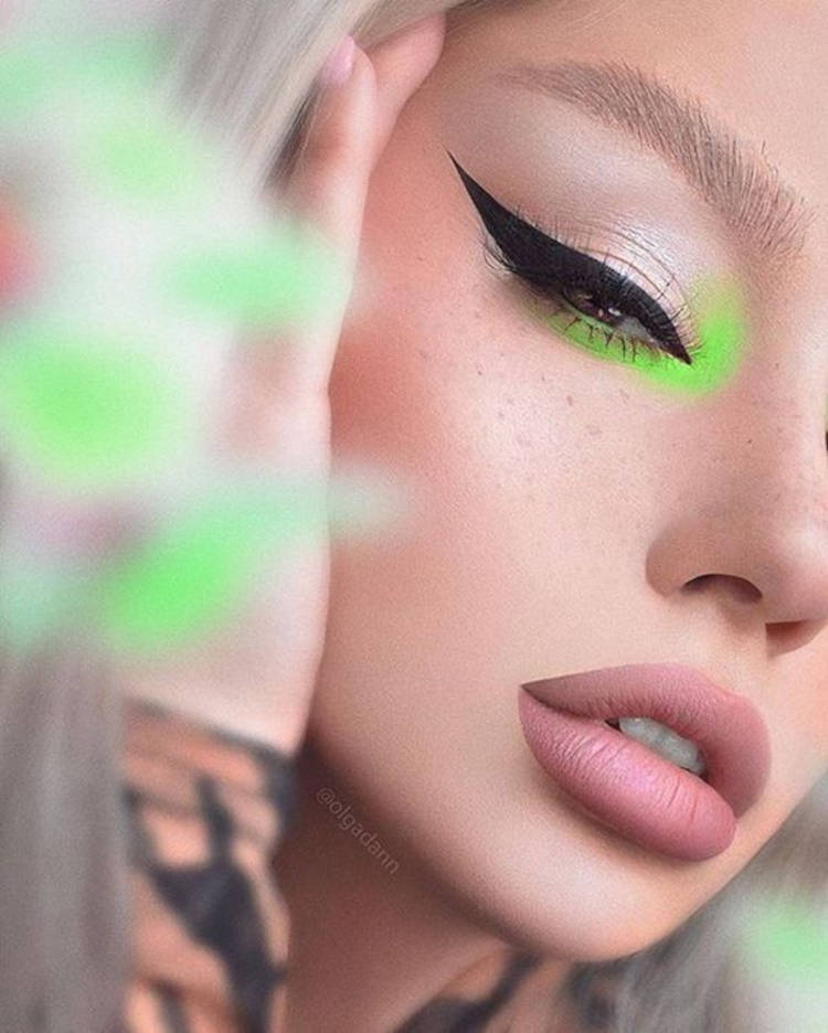 Zoom em pessoa de pele clara com batom matte rosa, delineado gatinho preto e canto o olho verde neon
