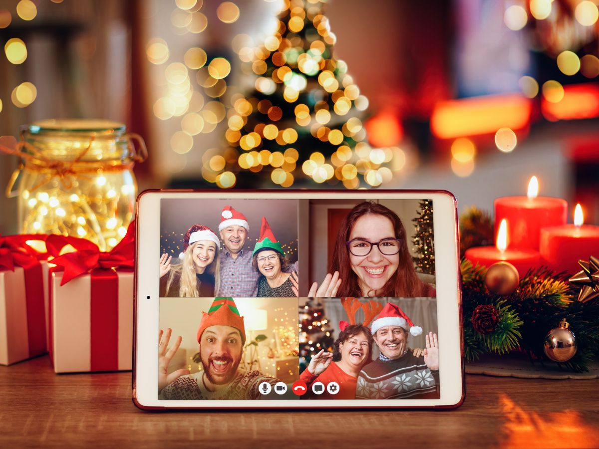 Foto de tablet com pessoas em chamada de vídeo. Fundo da imagem com decoração natalina, árvore, presentes e outros.