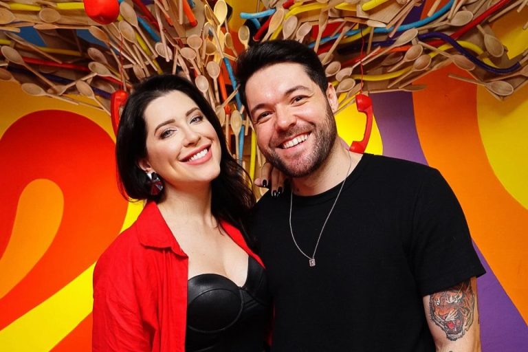 Andressa Ganacin de top preto e blazer vermelho abraçada com Nasser de camiseta preta, em fundo colorido