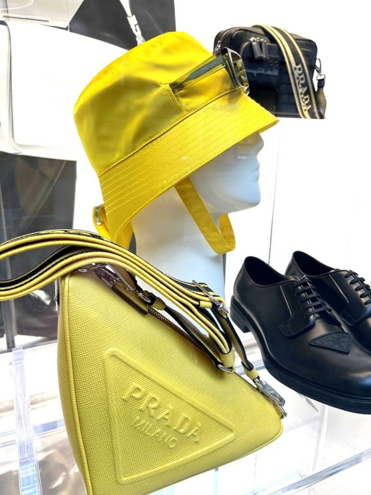 Manequim da marca Prada em Milão usando chapéu amarelo fluor, bolsa ao lado do manequim em amarelo fluor sapato e bolsa preata do lado direito direito da foto