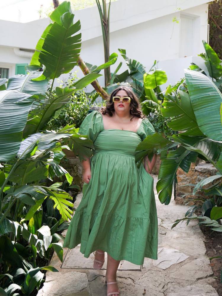 Mulher de pele clara e cabelo castanho usando óculos de sol e vestido romantico verde midi em meio à plantas