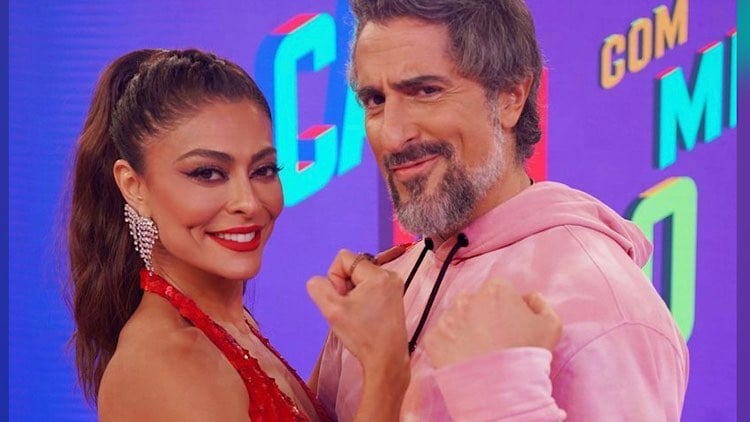 Juliana Paes posa ao lado de Marcos Mion no estúdio do Caldeirão, ela de vestido vermelho e ele de moletom rosa. Os dois sorriem e olha para câmera