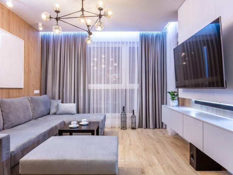 Sala pequena decorada com parede de madeira, lustre e sofá cinza.