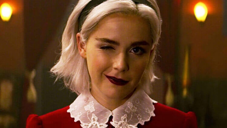 foto da personagem principal de O Mundo Sombrio de Sabrina. Ela usa tiara preta, vestido vermelho com gola branca e está piscando um olho
