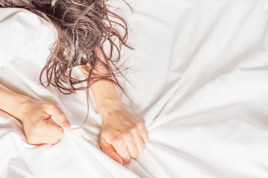 mulher deitada na cama com as duas mãos fechadas em punho agarrando o lençol branco