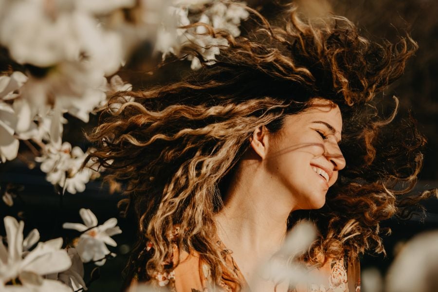 mulher de cabelos cacheados sorrindo ao lado de árvore com flores brancas