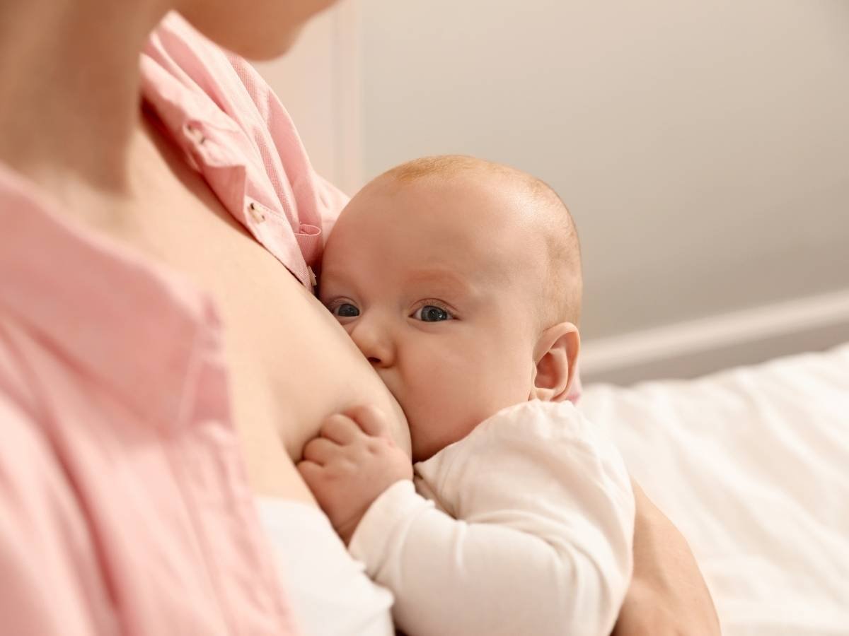 Mulher com camisa rosa aberta realizando a amamentação do seu bebê.