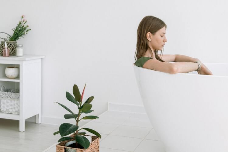 mulher em uma banheira branca com os olhos fechados e um vaso de planta ao redor
