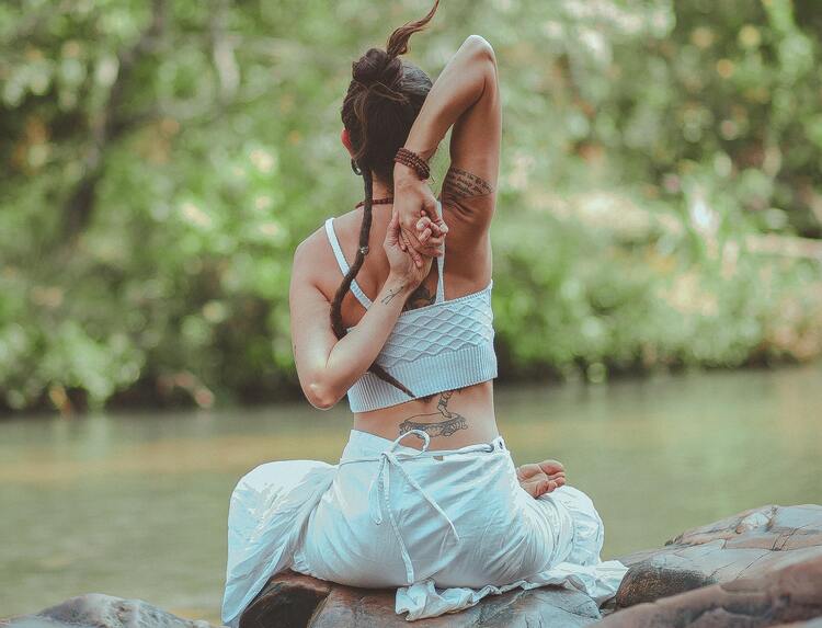 mulher usando cropped e saia brancos e fazendo posição de yoga na beira de um rio, filhos de Nanã