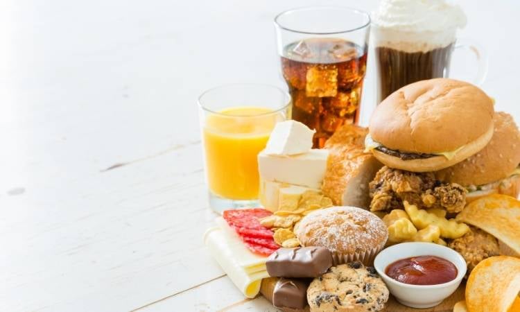 Mesa com comidas gordurosas: milkshake, refrigerante, hambúrguer, batata frita, ketchup, frios, chocolate, muffin e cookie
