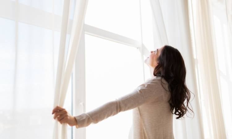 Mulher abrindo cortinas para evitar mofo da parede.