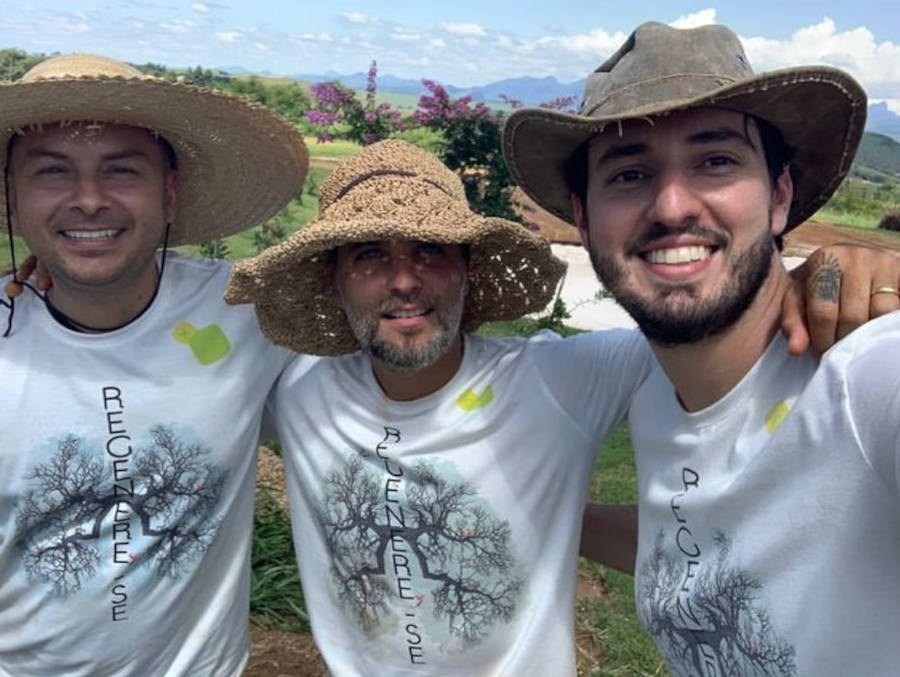 Imagem de Bruno Gagliasso no meio de dois amigos, os três estão com chapéus e blusa branc a escrito "regenere-se"