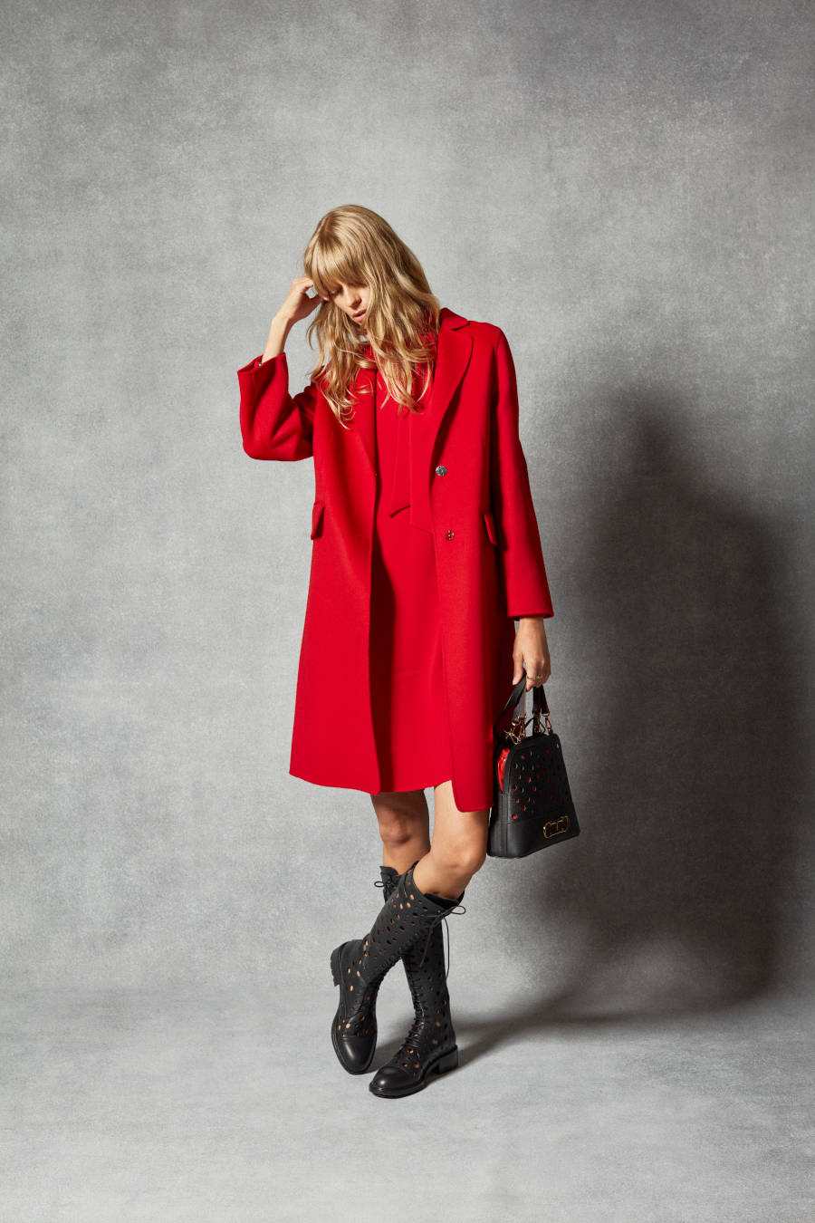 Foto de modelo usando bota de cano alto e sobretudo vermelho, fazendo contraste com a sua bolsa preta
