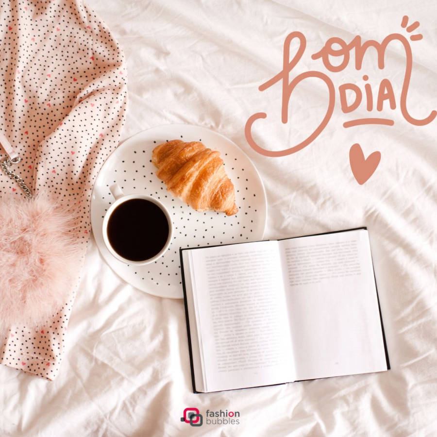 Imagem de um café da manhã com croissant e café ao lado de um caderno para anotações em cima de uma cama. Além disso, também possui a frase "Bom Dia" e um coração para a decoração