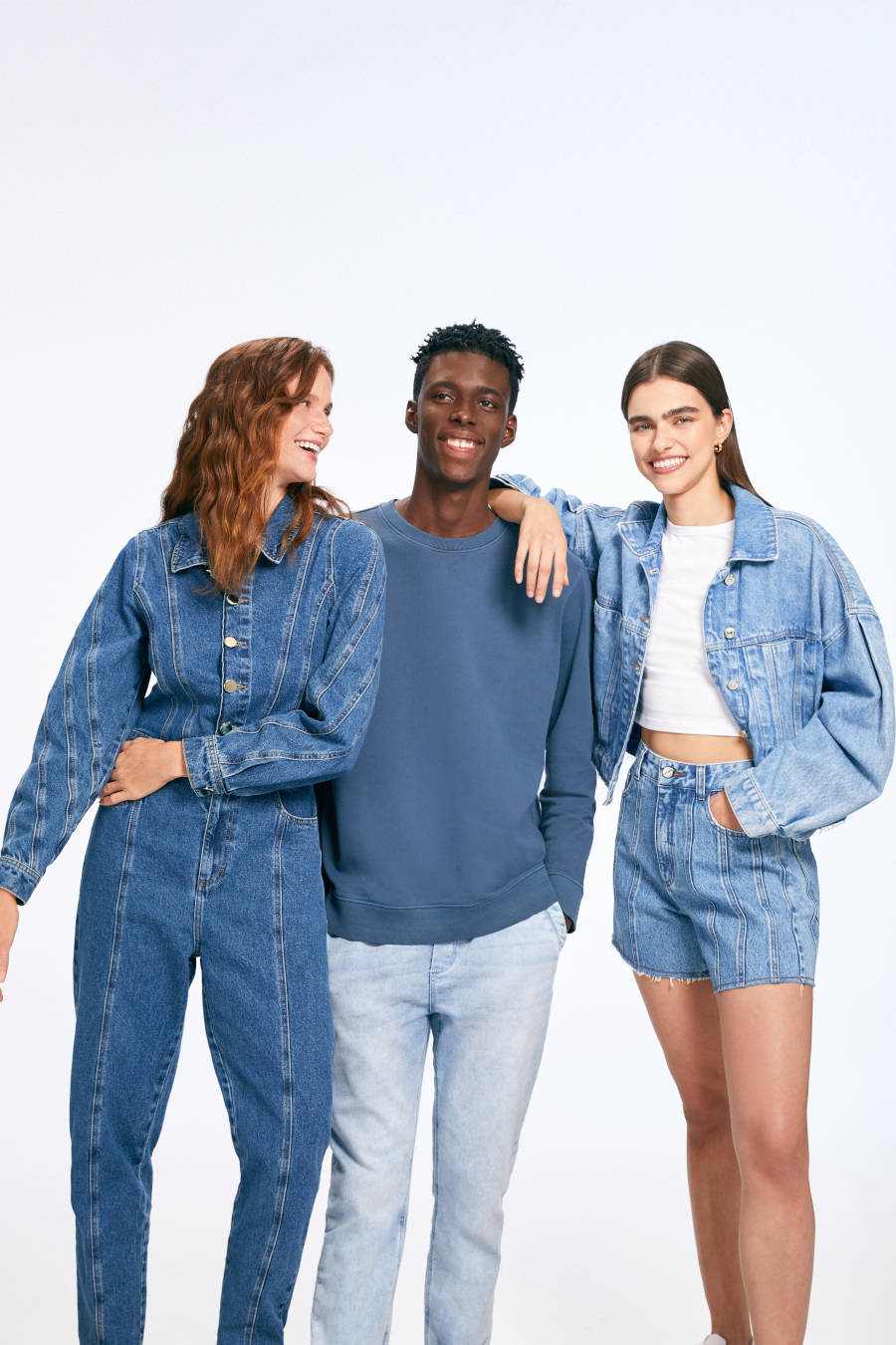 Imagem com três pessoas, duas mulheres, uma vestindo macacão jeans e outra short e jaquetas jeans e um homem vestindo calça jeans e blusa de manga azul