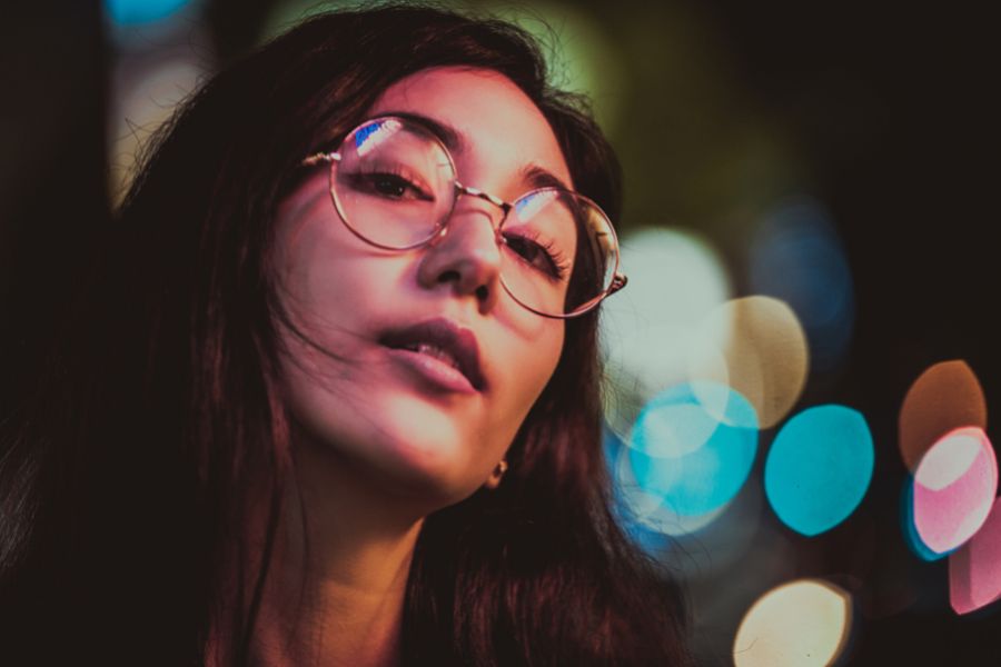 mulher de cabelos longos e castanhos usando armação redonda. Ao fundo, vemos luzes embaçadas