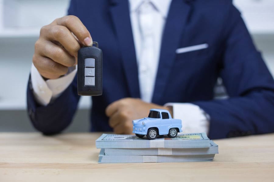 Imagem de um homem de terno segurando a chave de um carro, ao lado tem um carro de brinquedo azul em cima de uma pilha de dinheiro.