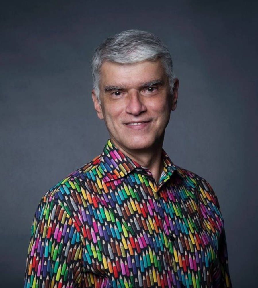 Foto do professor João Braga vestindo uma camisa com estampa de lápis coloridos e o fundo cinza