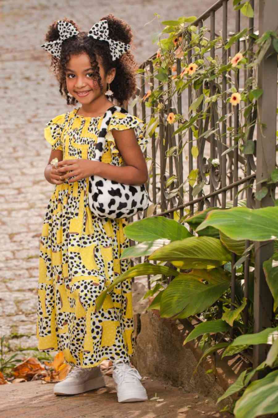 Foto de criança com vestido amarelo com estampas de animal, bolsa com estampa de vaquinha combinando com dois laços no cabelo com a mesma estampa