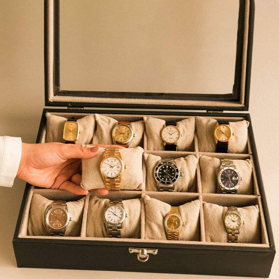 Imagem de caixa de relógios masculinos, com 11 relógios e um homem pegando um deles na mão