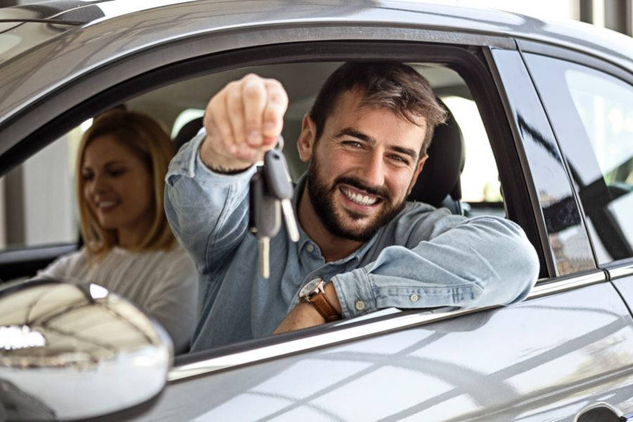 Foto de um homem com cabelo castanho e barba, vestindo uma amisa de manga longa jeans, dentro de um carro, no banco do motorista, e mostrando a chave do carro e sorrindo. Uma mulher loira sorrindo está sentada no banco do passageiro