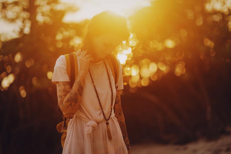 mulher tatuada, com roupas brancas e mochila usando japamala de Ho'oponopono e andando no sol